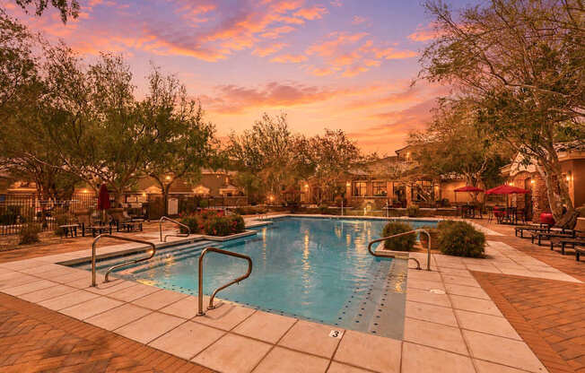 A swimming pool surrounded by trees and lounge chairs at sunset.