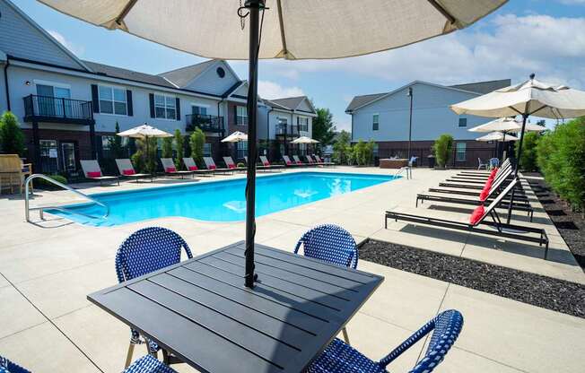 A table with chairs and an umbrella is in the foreground of a pool area.