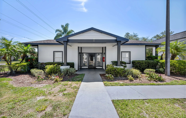 Charming white house with a welcoming entrance, surrounded by lush greenery and vibrant plants, under a clear blue sky.