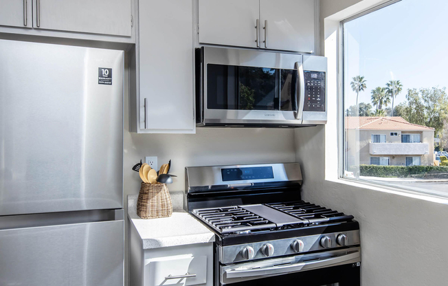 Modern kitchen featuring a stainless steel refrigerator, a microwave above the stove, and a gas range with five burners. Countertop appliances and utensils are neatly arranged, complemented by a window that shows a view of palm trees outside. The decor is bright and contemporary.