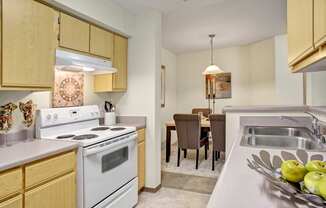 A functional kitchen here at Allegro featuring light wood cabinetry, neutral countertops, and a full appliance setup with a range and range hood, opening to a dining area with a rectangular table, upholstered chairs, and a hanging pendant light, while a double-basin sink and breakfast bar create a practical layout suited for everyday cooking and casual meals.