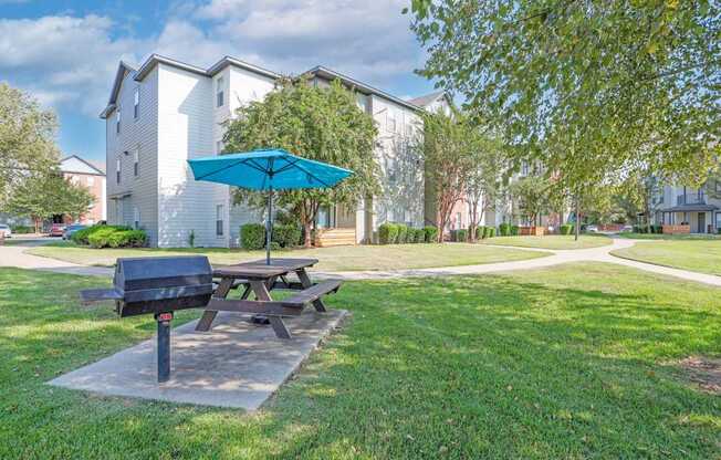 A picnic table with a blue umbrella is in the foreground of a grassy area with a building in the background.
