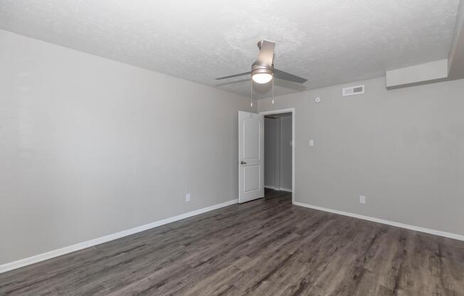 A spacious interior of an empty room featuring gray walls, a ceiling fan, and light-colored laminate flooring. The room has a doorway leading to another space, creating an open and airy feel. Natural light enhances the simplicity of the design, ideal for personalization.