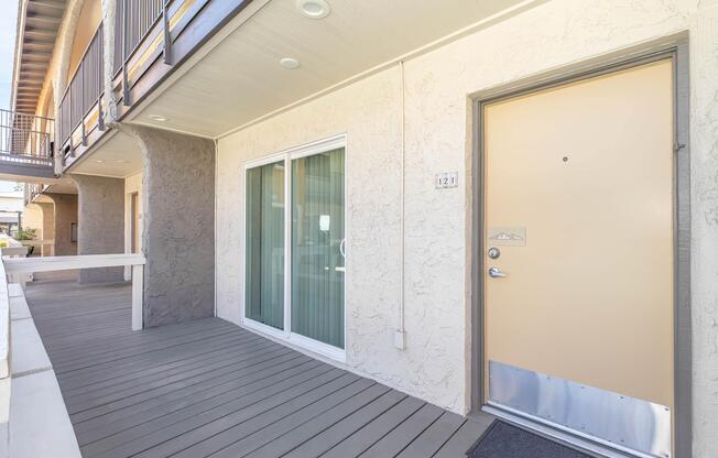 A front entrance of an apartment with a beige door, a light gray doormat, and sliding glass doors leading to an interior space. The building features a textured wall, wooden deck, and nearby balcony area. Natural light illuminates the entrance.