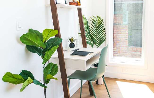 A room with a wooden floor, a green chair, a wooden ladder, a desk with a laptop, and a potted plant.