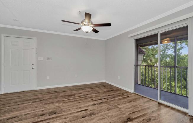 Interior view of a spacious room featuring a ceiling fan, light fixture, and wood-like flooring. A door leads to a balcony with sliding glass doors revealing a view of greenery outside. The walls are painted in a light gray color, creating a bright and inviting atmosphere.