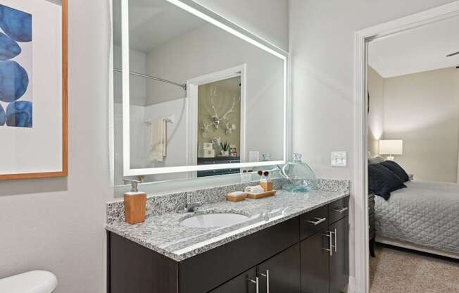 A bathroom with a sink, mirror, and a bed in the background.