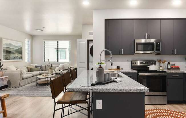 A modern open living space here at Reflect at Dobson Ranch showcasing a contemporary kitchen with dark flat-panel cabinetry, a spacious island with bar seating, polished countertops, and sleek appliances, opening into a comfortable living room with a large neutral sectional, layered rugs, soft natural light, and warm wood-style flooring.
