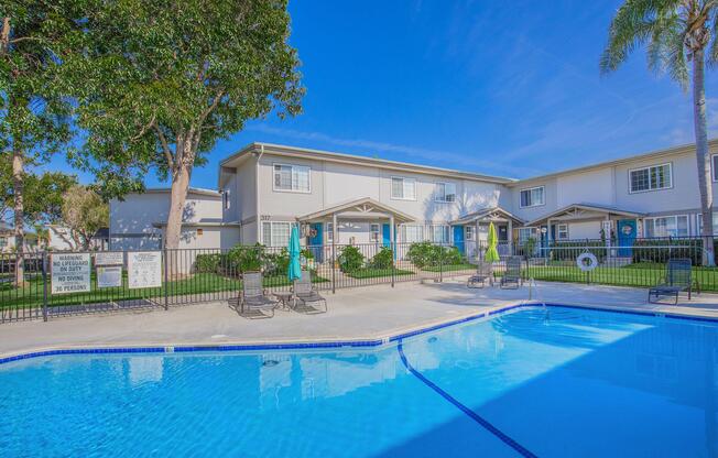A clear blue swimming pool with lounge chairs and umbrellas in a sunny courtyard. Surrounding the pool are green lawns and landscaped areas with trees. In the background, a two-story apartment building is visible against a bright blue sky. A sign near the pool indicates rules and safety information.