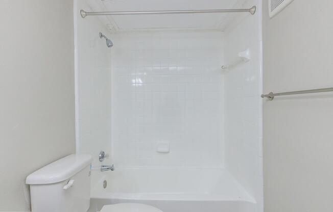A clean, minimalistic bathroom featuring a white bathtub with a showerhead, a white toilet, and a simple wall-mounted towel rack. The walls and tiles are light-colored, contributing to a bright and airy feel. The space is uncluttered and designed for practicality.