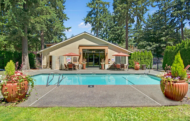 A landscaped outdoor swimming pool here at Asbury Park featuring a rectangular pool with metal handrails, a wide concrete pool deck, large decorative planters with greenery, shaded seating areas with patio tables and umbrellas, and a community building in the background, surrounded by mature trees and fencing for a peaceful, resort-style setting.