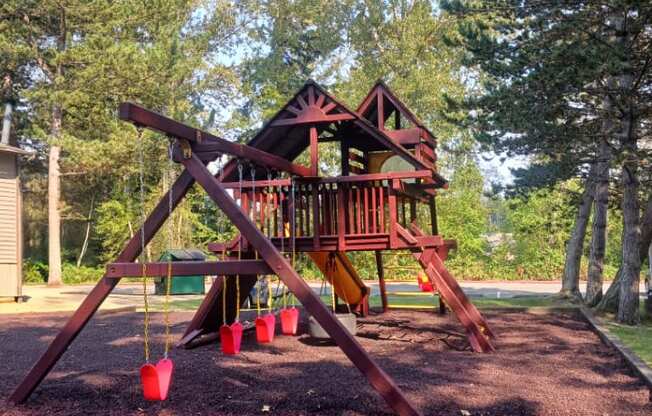 A wooden swing set with red swings is in the foreground of a park.