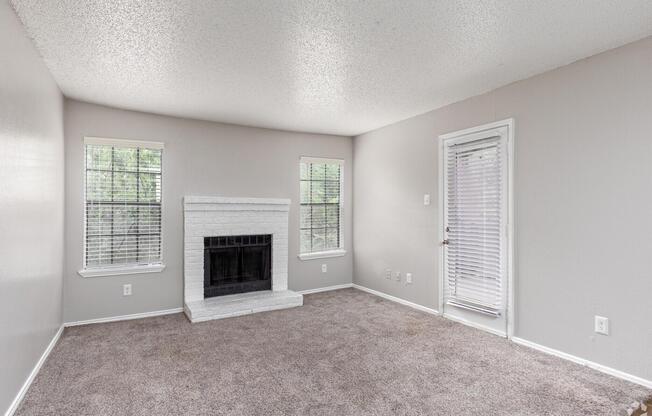 A spacious living room featuring light gray walls and carpet, a white brick fireplace as a focal point, and large windows allowing natural light. A door leads outside, and there are no furnishings, creating a minimalist and open atmosphere.