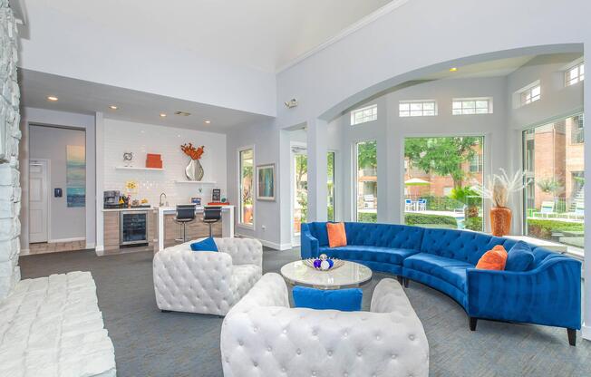 A bright and modern common area featuring a blue velvet sofa and two tufted white chairs surrounding a round table. The space includes large windows with a view of greenery outside, a kitchenette with sleek appliances, and decorative elements like artwork and plants.