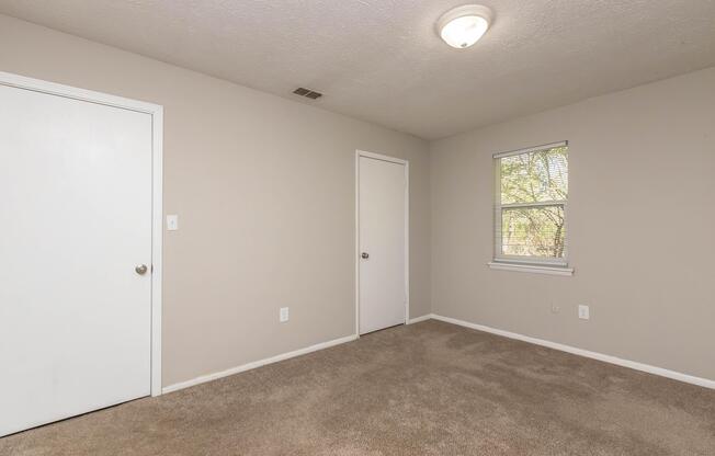 A simple, empty room featuring light gray walls and beige carpeting. A single window on the right allows natural light, while there are two doors: one visible in the left corner and another in the back wall. The ceiling light fixture provides overhead illumination, contributing to the bright and airy feel of the space.