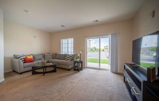 Image of living room, door leading to patio on the right, light tan painted walls, grey couch in left corner of room with coffee table in front.