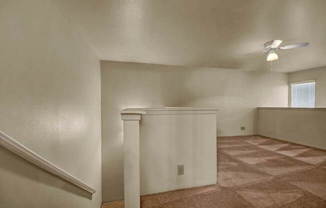 A spacious upstairs loft here at High Plains with soft beige carpet, neutral walls, and a ceiling fan with built-in lighting, featuring an open railing overlook and natural light from a window that creates a flexible, comfortable bonus living space.