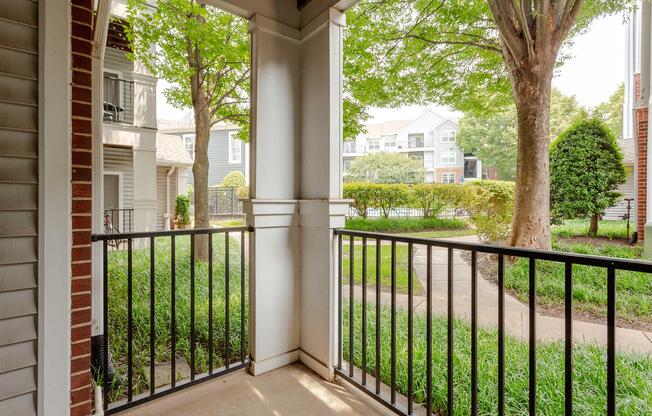View from a covered porch showcasing a green courtyard with manicured bushes and trees, surrounded by apartment buildings. The scene conveys a calm and welcoming atmosphere, ideal for relaxation and enjoying the outdoors. The porch features black railings and light-colored walls.