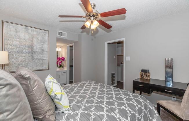 Cozy bedroom featuring a stylish gray and white patterned bedspread, decorative pillows, and a ceiling fan with wooden blades. A large piece of artwork hangs on the wall, and a bedside lamp adds warmth. A doorway leads to an adjacent room, enhancing the spacious feel of the area.