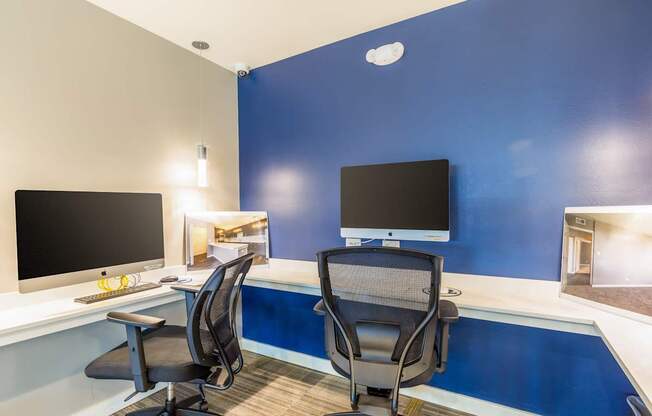 Two black computer monitors on a desk with two chairs.
