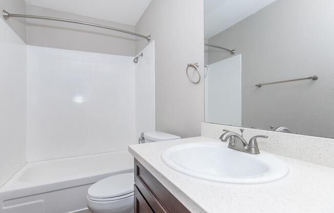 A clean bathroom featuring a white bathtub with an attached shower, a toilet, and a modern sink with a light-colored countertop. The walls are painted in a soft gray, and there is a mirror above the sink, along with a towel bar and a shower curtain rod. The overall design is simple and contemporary.