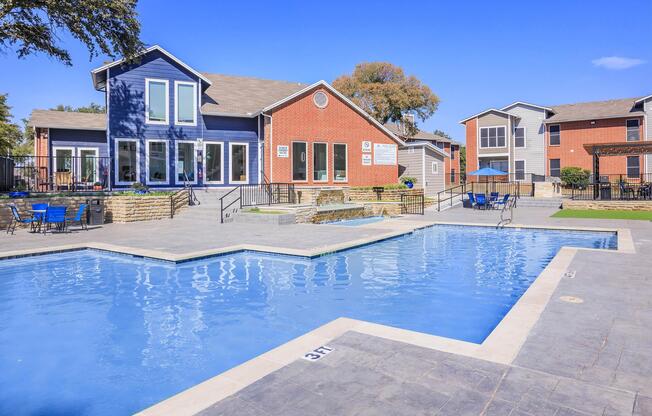 A clean, bright swimming pool with a surrounding patio area featuring blue lounge chairs. In the background, two colorful apartment buildings with a well-maintained landscape under a clear blue sky.