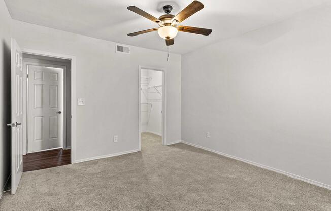Empty room with light gray walls and tan carpet. A ceiling fan with wooden blades hangs from the ceiling. Two doors are visible, one leading to a closet and the other to a hallway. Natural light is minimal, creating a simple and clean aesthetic. No furniture or decor present.
