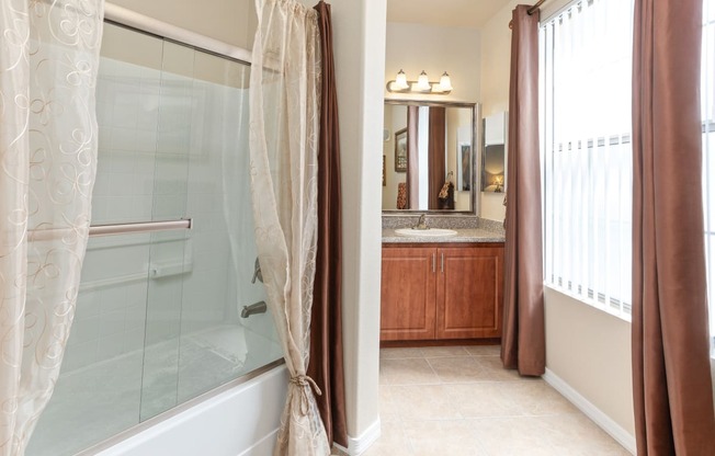 Master Bathroom showing double sink vanity in the background with a large framed mirror and step in tub with glass doors and accent curtains.