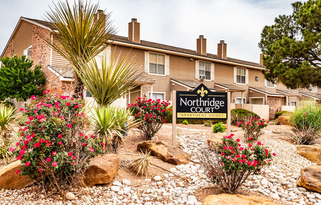 A landscaped monument sign here at Northridge Court featuring the Northridge Court Apartment Homes signage surrounded by colorful flowering shrubs, desert-style plants, decorative rocks, and mature greenery, with classic two-story apartment buildings and neutral siding creating a welcoming community entrance backdrop.