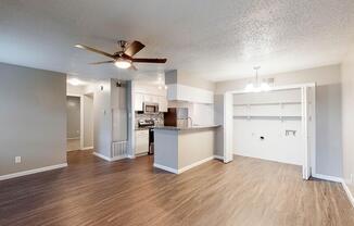 A spacious, modern kitchen and living area featuring a ceiling fan, stainless steel appliances, a breakfast bar, and light wood flooring. The room is well-lit with natural light, showcasing a neutral color palette and an open layout. A small closet is visible on the right side.