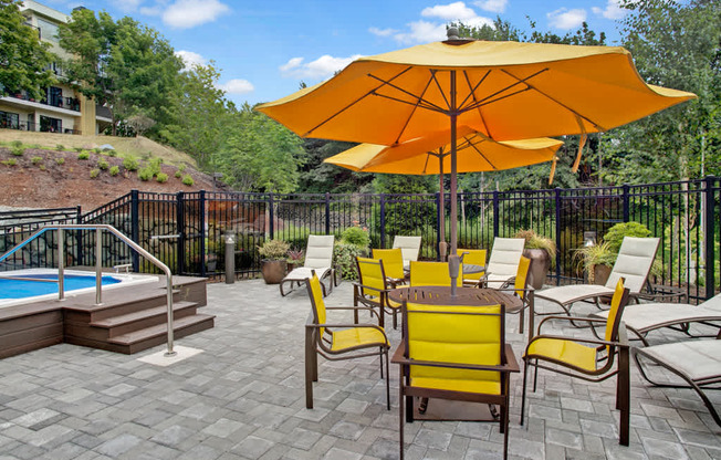 A resort-style outdoor patio here at Heritage Hills featuring a paved seating area with yellow cushioned chairs arranged around café tables beneath large umbrellas. The space includes nearby pool and spa access, surrounding lounge chairs, landscaped greenery, and black metal fencing, creating a relaxing setting for outdoor gatherings and leisure.