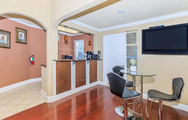 Modern kitchen space featuring a bar area with a glass table and black stools. The walls are painted in warm tones, with framed artwork and a fire extinguisher visible. A microwave and coffee maker are on the counter, and a flat-screen TV is mounted nearby. The flooring is wooden, creating a cozy atmosphere.