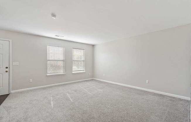 A room with a carpeted floor, two windows with blinds, and a door.