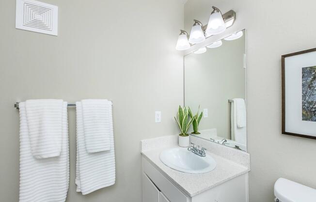 A clean bathroom featuring a white countertop sink with a modern faucet, an illuminated mirror above the sink, two neatly hung white towels, and a small potted plant. The walls are painted in a light beige color, creating a bright and airy atmosphere. A decorative framed artwork is also visible.