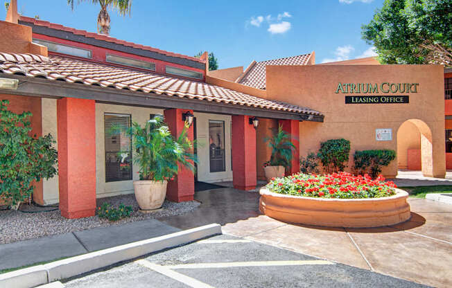 A welcoming leasing office exterior here at Aspire Pinnacle Park featuring warm Southwestern architecture with stucco walls, red accent columns, and a tile roof. Lush landscaping with palm trees and a vibrant flower bed frames the covered entry, while clear signage and a shaded walkway create an inviting first impression for residents and guests.