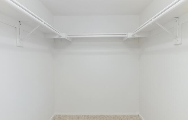 Empty room with white walls and a beige carpeted floor featuring two white closet rods mounted on brackets. The space is devoid of any furniture or items, creating a minimalistic and open appearance.