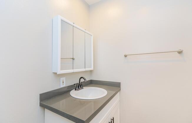 A modern bathroom sink area featuring a circular basin on a gray countertop, a white cabinet below, and a mirror with three panels above. The wall is a soft light color, and there is a towel bar mounted to the side.