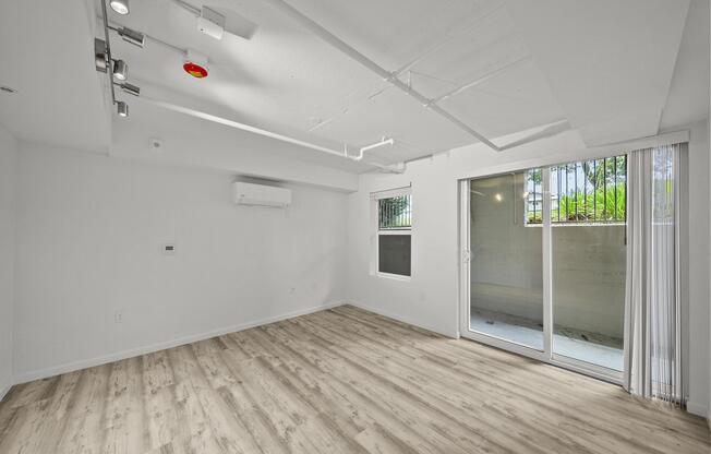 A spacious, empty room featuring light-colored walls and wooden flooring. There is an air conditioning unit on the wall, track lighting on the ceiling, and large glass sliding doors that lead to a small outdoor area. The room is bright with natural light flowing in.