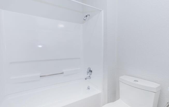 A clean, modern bathroom featuring a white bathtub with a showerhead, a silver grab bar, and a white toilet. The walls are painted a light color, creating a bright and spacious atmosphere.