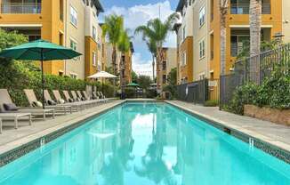 A swimming pool surrounded by lounge chairs and umbrellas in front of apartment buildings.