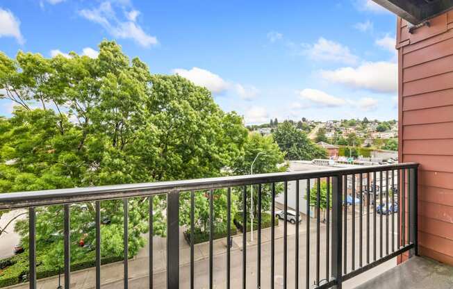 A balcony with a black railing overlooks a green tree and a street.