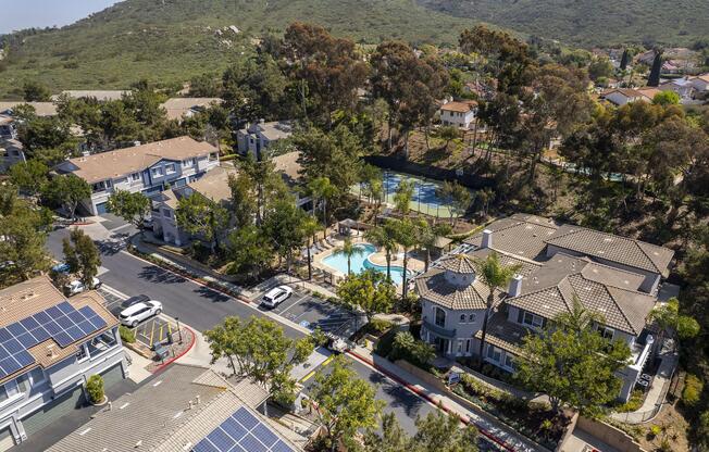 Aerial view of a suburban neighborhood featuring multiple residential buildings with lush landscaping, a swimming pool, and tennis courts. Solar panels are visible on rooftops, and the area is surrounded by green hills. The scene conveys a peaceful and well-maintained community atmosphere.