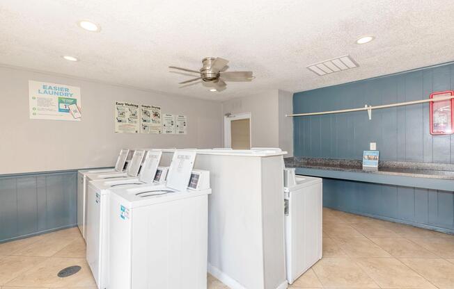 A laundry room featuring several white washing machines lined up against a wall. There is a countertop for folding laundry, a ceiling fan, and a bulletin board with laundry instructions or guidelines. The walls are painted a light blue, and the space appears bright and clean.