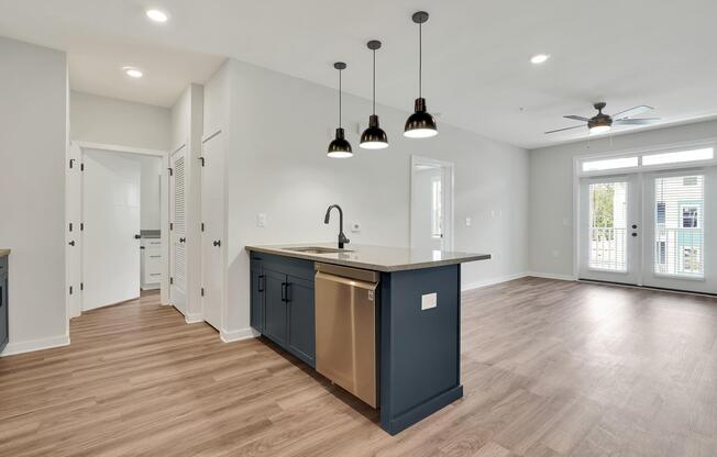 Modern kitchen and living area featuring a central island with a stainless steel sink and dishwasher. Pendant lights hang above the island. The space includes light wood flooring, white walls, and a door leading to a patio. Additional doors can be seen in the background. Bright and airy atmosphere.