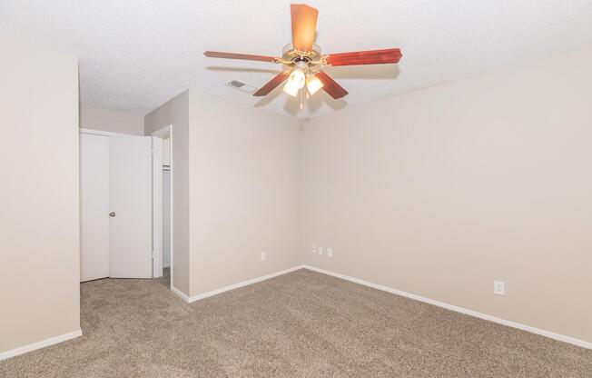 A spacious room featuring light beige walls and plush carpet. A ceiling fan with wooden blades is mounted in the center, providing ventilation. To one side, there is a closet with a white door, and another door leads outside of the room. The overall atmosphere is bright and inviting, perfect for various uses.