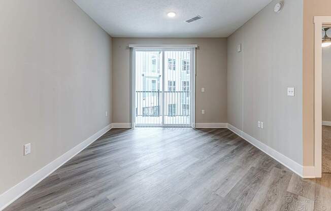 A spacious room with light wood flooring and a view of a balcony through a glass door.