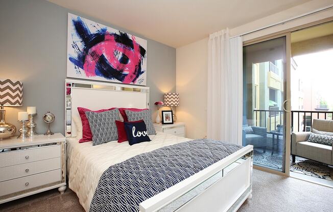 A bedroom with a large bed, a painting above it, and a chair in the doorway at The Kitt at Warner Center Apartments, Woodland Hills, CA