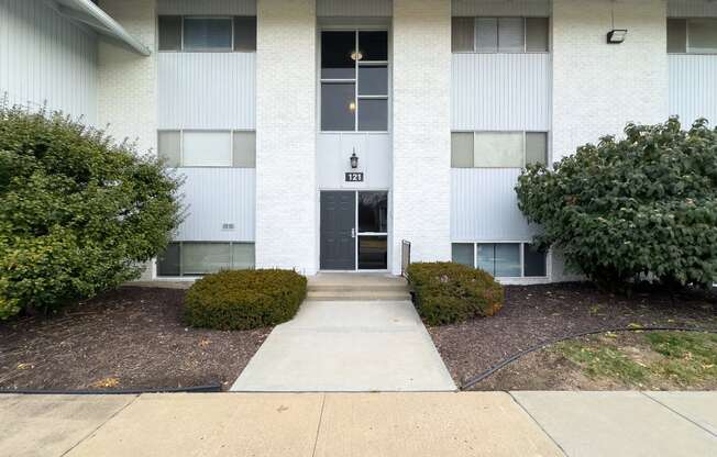 the front entrance to a white building with bushes and a sidewalk