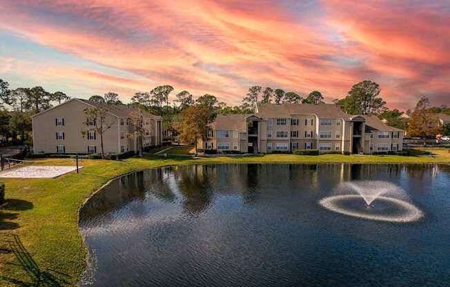 A sunset view of a lake with a fountain in the middle and apartment buildings in the background.