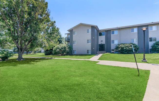 Apartment building with multiple floors, surrounded by green grass and trees. A pathway leads to the entrance, and a lamppost stands nearby. The sky is clear and blue, contributing to a bright, inviting atmosphere.